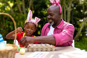 girl and grandfather wearing bunny ears while painting eggs on easter day