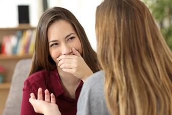 Ashamed woman hiding her smile in a conversation with a friend sitting on a couch at home